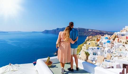 Couple standing on white terrace overlooking Santorini's blue Aegean Sea, caldera cliffs, and whitewashed village buildings